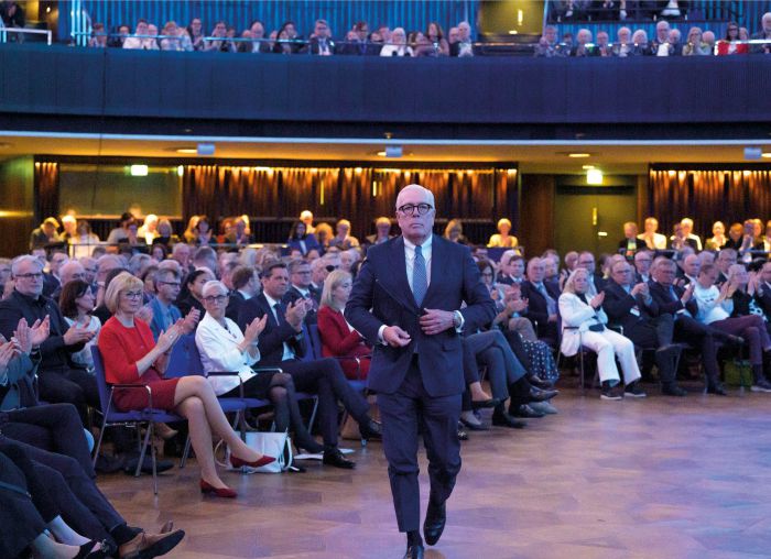 Sitzung der Ärztekammer mit zahlreichen Teilnehmern an Tischen, Vortragendem am Podium und Präsentation im Konferenzraum.