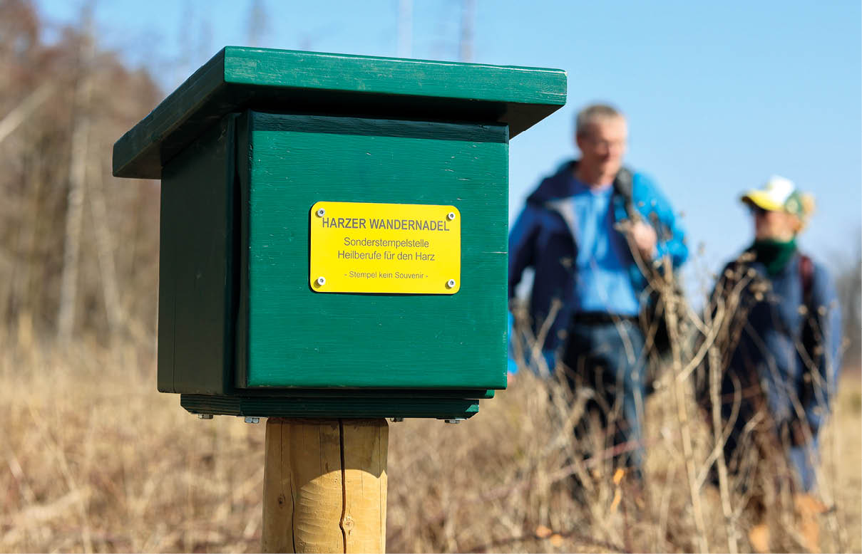 Harzer Wandernadel Stempelstelle für Heilberufe, im Hintergrund zwei wandernde Personen