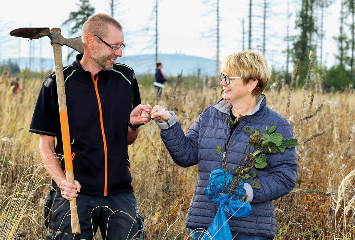 Zwei Personen beim Bäumepflanzen im Freien, lächelnd mit Faustgruß, eine Person hält Setzlinge