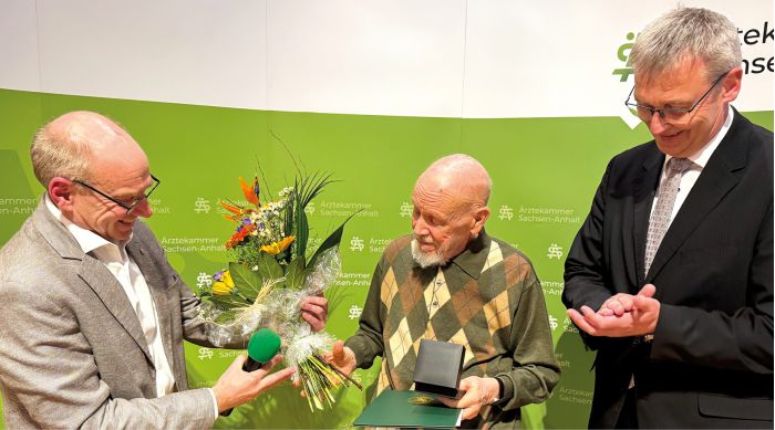 Sitzung der Ärztekammer mit zahlreichen Teilnehmern an Tischen, Vortragendem am Podium und Präsentation im Konferenzraum.
