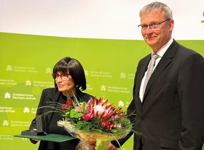 Sitzung der Ärztekammer mit zahlreichen Teilnehmern an Tischen, Vortragendem am Podium und Präsentation im Konferenzraum.