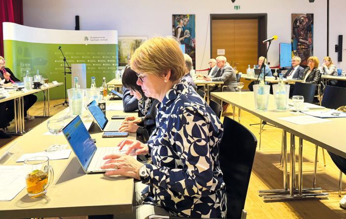 Sitzung der Ärztekammer mit zahlreichen Teilnehmern an Tischen, Vortragendem am Podium und Präsentation im Konferenzraum.