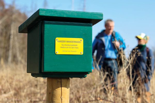 Harzer Wandernadel Stempelstelle für Heilberufe, im Hintergrund zwei wandernde Personen