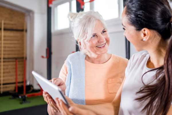 Ältere Frau spricht nach dem Training lächelnd mit einer Trainerin, die ihr Informationen auf einem Tablet zeigt.