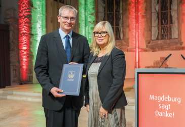 Ein Mann erhält eine Auszeichnung im Rathaus Magdeburg, steht mit einer Frau vor beleuchteter Kulisse und einem Pult mit der Aufschrift „Magdeburg sagt Danke!“.