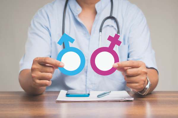 Arzt hält Symbole für männlich und weiblich in den Händen; Fokus auf Geschlechterzeichen vor medizinischem Hintergrund.