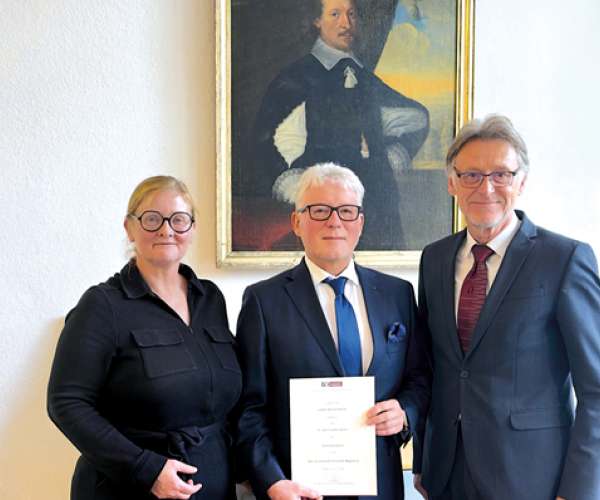 Drei Personen stehen vor einem historischen Porträt; der Mann in der Mitte hält eine offizielle Urkunde in den Händen.