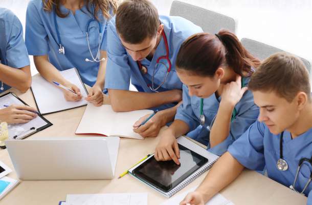 Medizinstudierende in blauen Kitteln lernen gemeinsam mit Tablet, Laptop und Unterlagen in der Ausbildung.