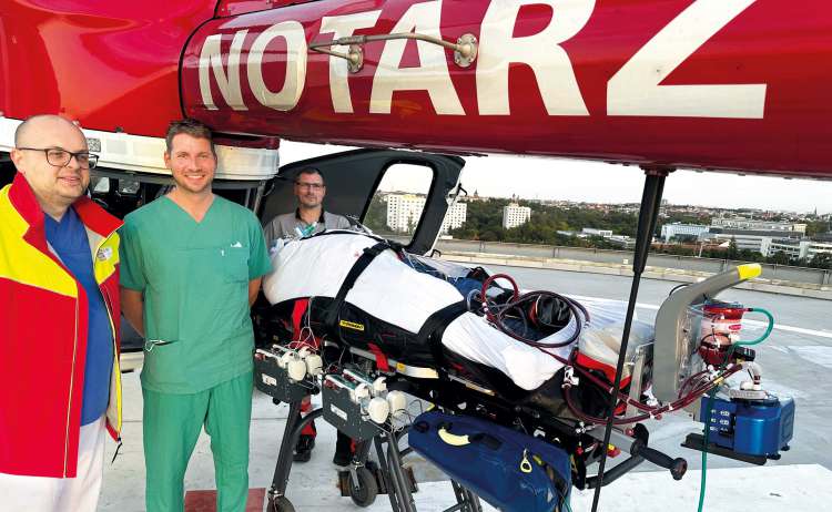 Notarztteam mit Patient auf Trage und ECMO-System vor Rettungshubschrauber auf dem Dachlandeplatz einer Klinik.