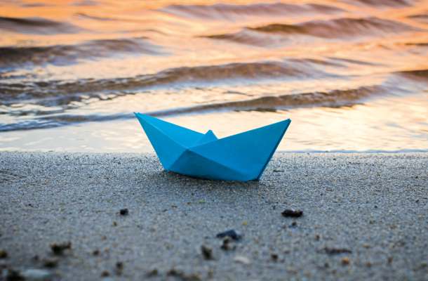 Blaues Papierboot liegt am Strand vor sanften Wellen im Abendlicht – Symbolbild für Erinnerung, Hoffnung und Abschied.