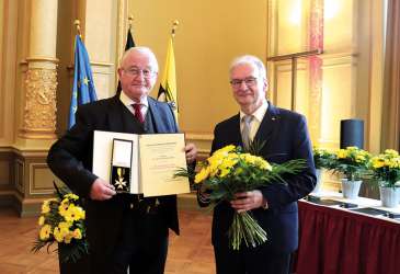 Zwei Männer bei einer Ehrung; einer hält Urkunde und Orden, der andere einen Blumenstrauß, im festlichen Saal mit Flaggen.