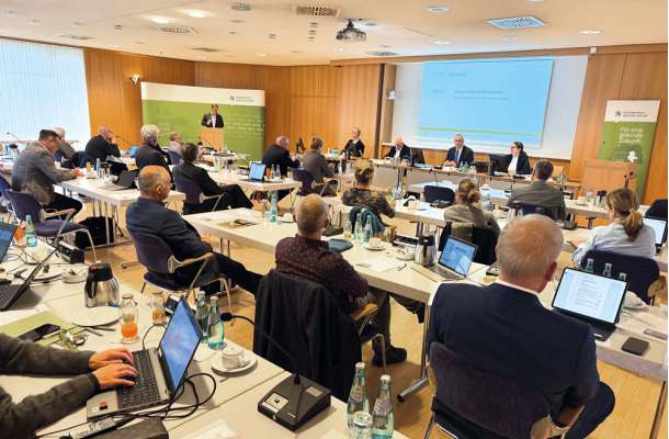 Sitzung der Ärztekammer mit zahlreichen Teilnehmern an Tischen, Vortragendem am Podium und Präsentation im Konferenzraum.