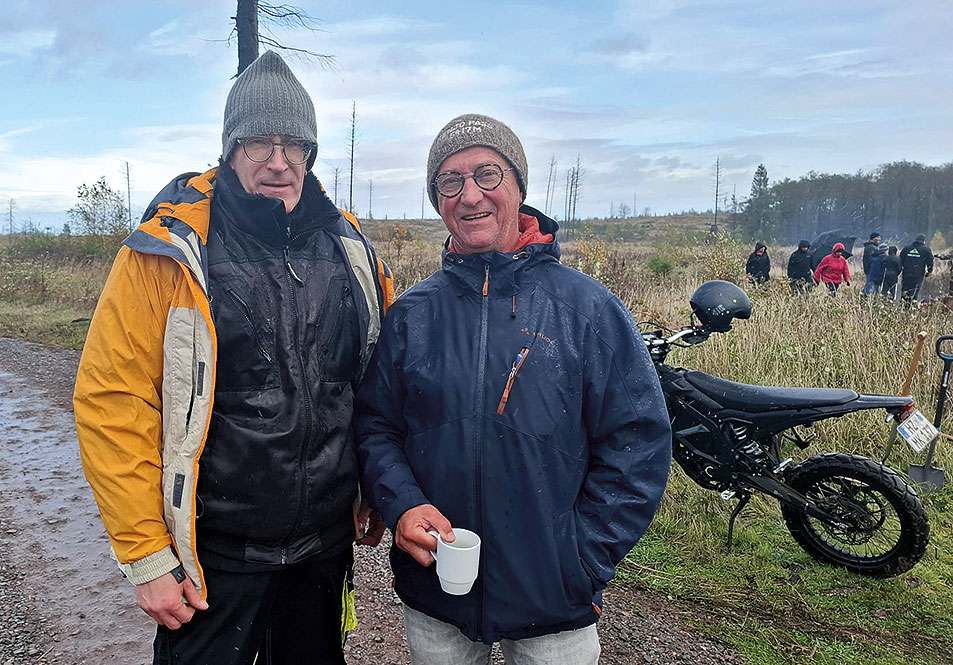 Zwei Männer in Regenkleidung stehen bei einer Pflanzaktion im Freien; einer hält eine Tasse, im Hintergrund Helfer und ein Motorrad.