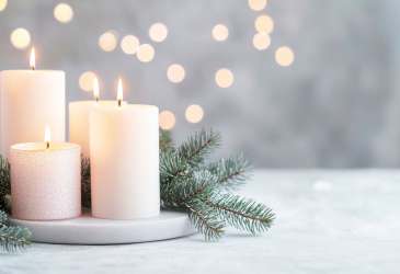 Vier brennende weiße Kerzen mit Tannenzweigen auf heller Fläche, unscharfe Lichtpunkte im Hintergrund sorgen für festliche Stimmung.