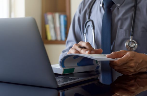 Arzt mit Stethoskop liest Fachliteratur am Schreibtisch neben einem geöffneten Laptop.