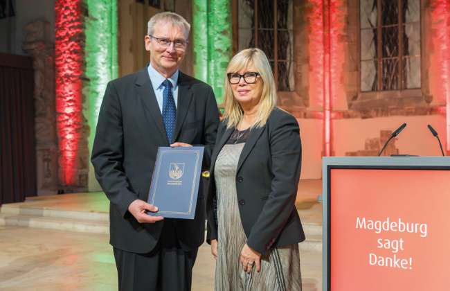 Ein Mann erhält eine Auszeichnung im Rathaus Magdeburg, steht mit einer Frau vor beleuchteter Kulisse und einem Pult mit der Aufschrift „Magdeburg sagt Danke!“.