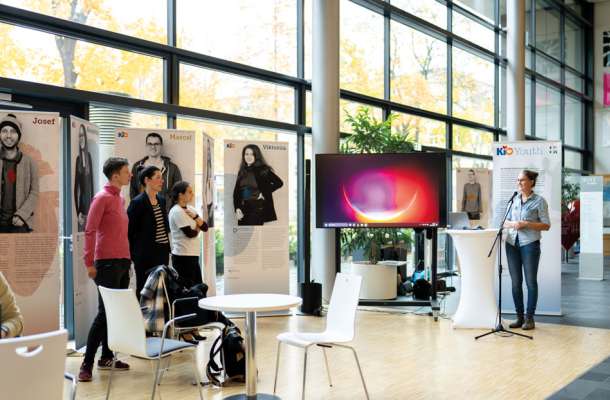 Ausstellung mit Jugendporträts und Vortragender am Mikrofon in einem lichtdurchfluteten Raum mit großer Fensterfront.