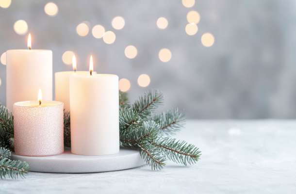 Vier brennende weiße Kerzen mit Tannenzweigen auf heller Fläche, unscharfe Lichtpunkte im Hintergrund sorgen für festliche Stimmung.