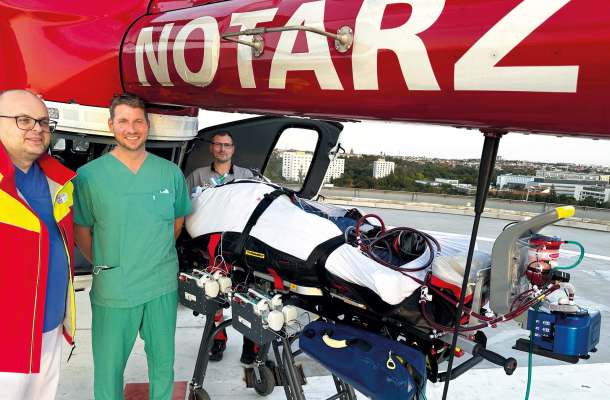 Notarztteam mit Patient auf Trage und ECMO-System vor Rettungshubschrauber auf dem Dachlandeplatz einer Klinik.