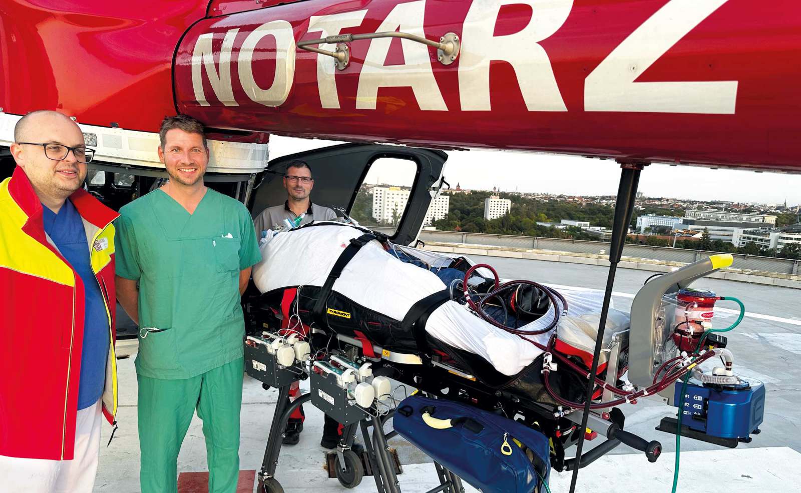 Notarztteam mit Patient auf Trage und ECMO-System vor Rettungshubschrauber auf dem Dachlandeplatz einer Klinik.