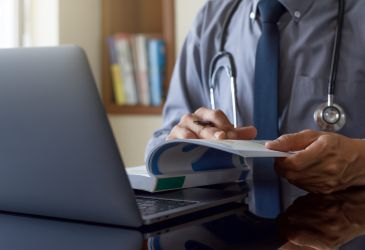 Arzt mit Stethoskop liest Fachliteratur am Schreibtisch neben einem geöffneten Laptop.