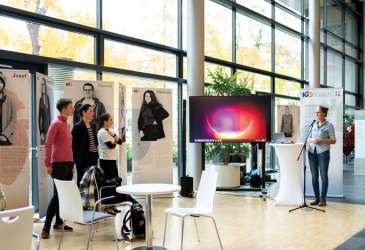 Ausstellung mit Jugendporträts und Vortragender am Mikrofon in einem lichtdurchfluteten Raum mit großer Fensterfront.