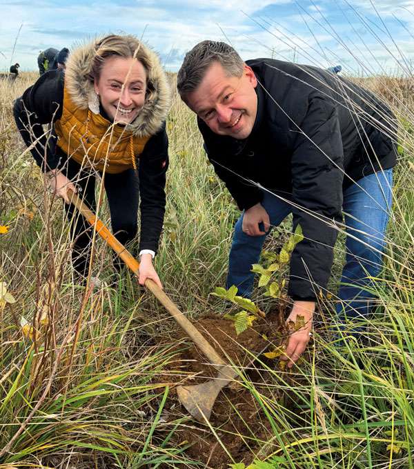 Zwei Personen pflanzen gemeinsam einen jungen Baum im hohen Gras und lächeln dabei in die Kamera.