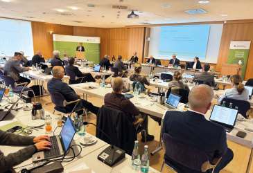 Sitzung der Ärztekammer mit zahlreichen Teilnehmern an Tischen, Vortragendem am Podium und Präsentation im Konferenzraum.