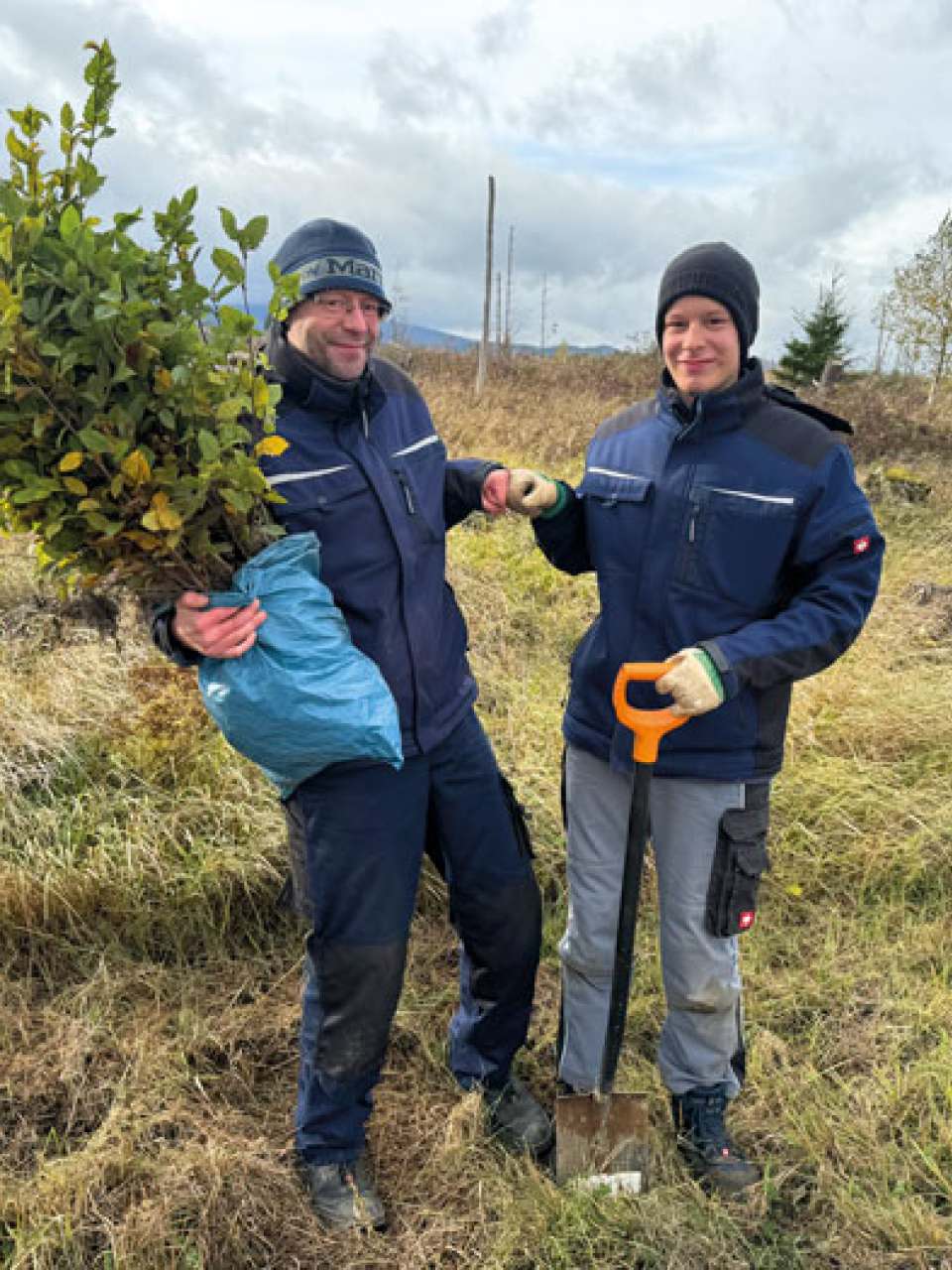 Zwei Helfer bei einer Pflanzaktion, einer hält junge Setzlinge, der andere stützt sich auf einen Spaten; beide tragen Outdoorkleidung.