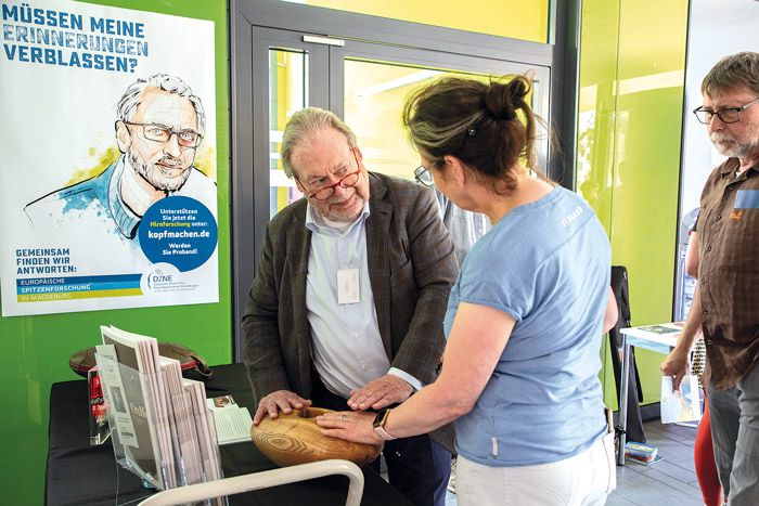 Besucher informiert sich beim Ethiktag an einem Stand zur Demenzforschung und testet ein haptisches Holzmodell im Gespräch mit Fachleuten.