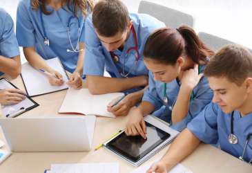 Medizinstudierende in blauen Kitteln lernen gemeinsam mit Tablet, Laptop und Unterlagen in der Ausbildung.