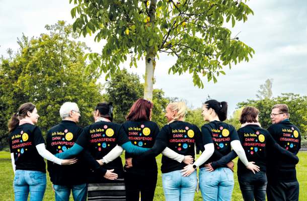 Gruppe von Organempfängern mit Shirts „Ich lebe dank Organspende“ steht vereint unter einem Baum im Park.