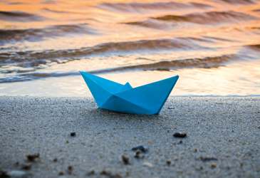 Blaues Papierboot liegt am Strand vor sanften Wellen im Abendlicht – Symbolbild für Erinnerung, Hoffnung und Abschied.