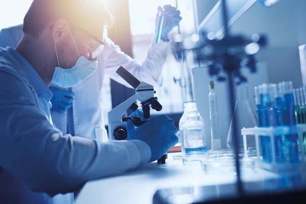 Forscher im Labor arbeitet am Mikroskop, umgeben von Proben und Reagenzgläsern; ein Kollege hält eine blaue Flüssigkeit im Testrohr.